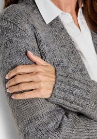 Eine Frau mit verschränkten Armen trägt einen grauen Strickpullover mit einem weißen Hemd darunter.