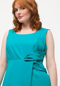 Abito senza maniche in teal con tessuto liscio, scollatura quadrata e un dettaglio decorativo a nodo in vita, che offre una silhouette moderna.