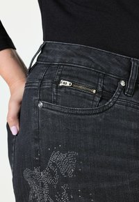 Jeans de mezclilla negra con un bolsillo con cremallera, ajuste estructurado y un patrón sutil de rhinestones en el costado. Textura suave con acentos plateados.