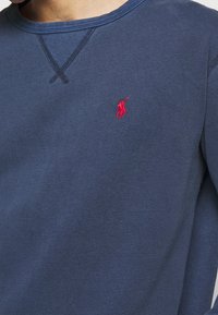 Blå crewneck sweatshirt med en liten röd broderad polo-spelare logga på vänster bröst och förstärkt sömnad nära kragen.