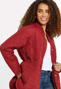 Giacca imbottita rossa con colletto arrotondato, vita elasticizzata e bottoni a pressione. Presenta una texture del tessuto liscia e curve sottili nel motivo.