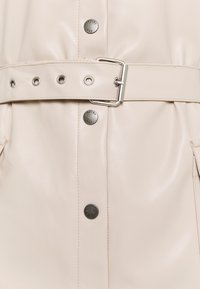 Beige konstläderjacka med bälte i midjan, tryckknappar och sidfickor, med en slät yta och minimalistisk design.