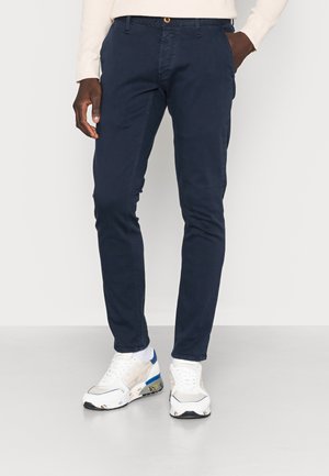 Mann trägt marineblaue, schmal geschnittene Hose, weiße Sneakers mit blauen Akzenten und ein hellbeiges Langarmshirt, steht vor einem schlichten Hintergrund.