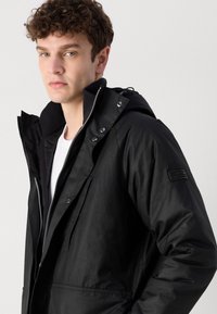 Fekete vízálló dzseki kapucnival, cipzáras záródással, patentgombokkal és elülső zsebekkel. Jellegzetes textúrájú gallérral és logós folttal a ujján.