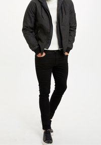 Sort bomberjakke med lynlås, ribbedemanchetter, to sidelommer, parret med slim-fit sorte jeans og sorte snørebåndssko. Hvid trøje indeni.