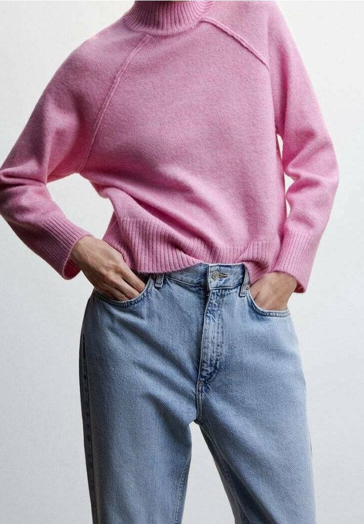 Personne portant un pull à col roulé rose rentré dans un jean taille haute bleu clair, les mains dans les poches, sur un fond uni.