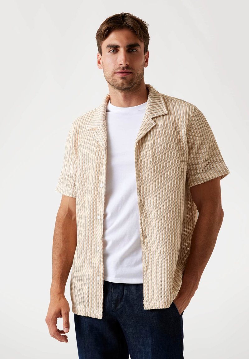 Short-sleeve, beige shirt with vertical stripes, a camp collar, and buttons down the front worn over a white t-shirt, paired with dark jeans.