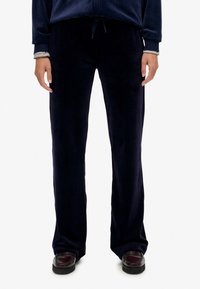 Persoon in zwarte velours wijdbeenbroek met strikkoord in de taille en donkerbruine instapschoenen, staande tegen een effen achtergrond.