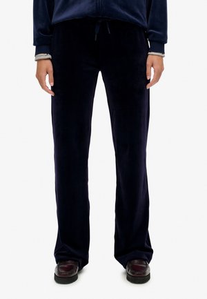 Person iført sorte velour brede bukser med snoretræk i taljen og mørkebrune loafers, stående mod en ensfarvet baggrund.