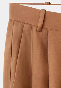Close-up of brown pleated trousers waistband with belt loop and textured fabric.
