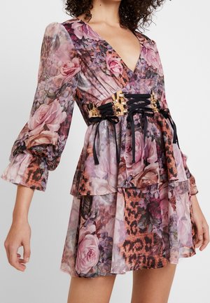 Femme portant une robe superposée à imprimé floral et léopard, avec des manches bouffantes en tulle transparent et une ceinture noire à lacets ornée d'accents dorés.