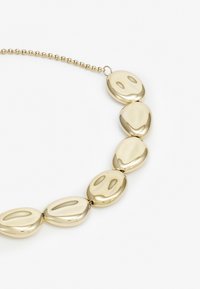 Zlatý náhrdelník s hladkými oválnými články s jemnými prohlubněmi, připevněný k jemnému řetízku pro bezpečné nošení.