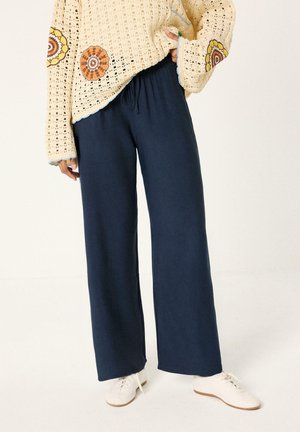 Femme portant un pull en crochet crème avec des motifs floraux orange, un pantalon large bleu marine et des chaussures blanches à lacets, debout la main dans la poche.