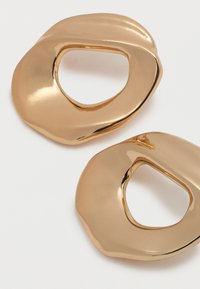 Δύο λάμποντα χρυσά μεταλλικά δαχτυλίδια ακανόνιστου σχήματος με ανοικτά κέντρα σε λευκό φόντο.