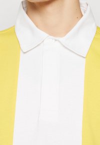 Collared polo shirt featuring a white front panel and yellow side panels, made of a smooth cotton blend material. Three-button placket.