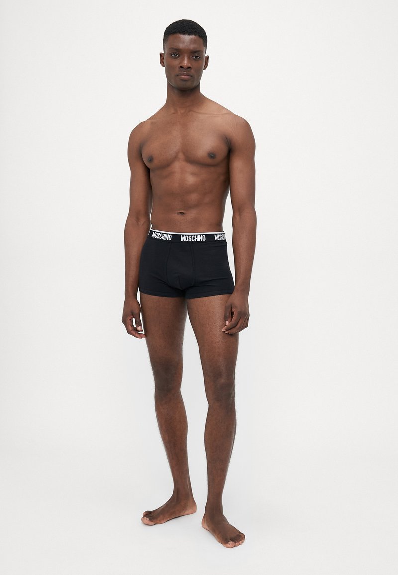 Black cotton boxer shorts with a white "MOSCHINO" logo on the waistband, modeled by a person standing barefoot against a plain background.