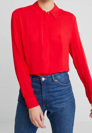Personne portant une chemise rouge vif à manches longues boutonnée, rentrée dans un jean en denim bleu taille haute, debout devant un fond uni.
