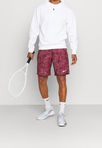 Sweat à capuche blanc avec poche avant, short bordeaux à motifs, baskets blanches, tenant une raquette de tennis blanche. Logos Nike sur le short et les chaussures.