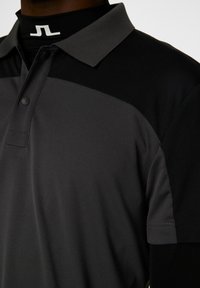 Herrpolo i svart och mörkgrått, tillverkad av andningsbart material. Har en krage och en kortärmad design med knappslå.