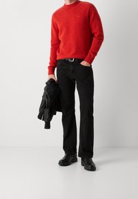 Roter Strickpullover mit gerippten Bündchen, kombiniert mit schwarzen Jeans in geradem Schnitt. Hält eine schwarze Lederjacke. Schwarze Stiefel vervollständigen das Outfit.