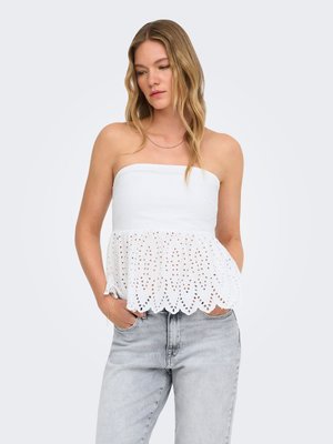 Femme aux longs cheveux blonds portant un top blanc sans bretelles en dentelle ajourée et un jean gris clair, mains dans les poches, regardant vers le bas.
