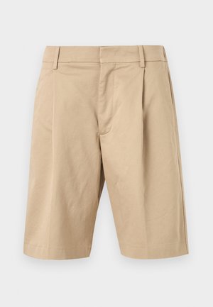 Ung mann står med hendene i lommene, iført en lyseblå kortermet skjorte med knapper, beige knelang shorts og svarte sandaler.