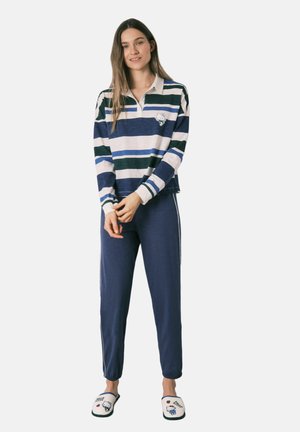 Kvinde står iført langærmet stribet polo-shirt, marineblå bukser og hvide Hello Kitty-hjemmesko mod en ensfarvet baggrund.