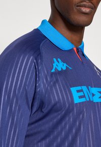 Polo blu navy con colletto azzurro, righe verticali e dettagli del logo in blu. Il tessuto sembra liscio e leggero.