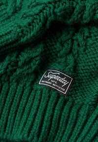 Vihreä neulottu pipo, jossa on ribbitekstuuri. Pipossa on musta etiketti, jossa lukee "Superdry" ja "Outdoor Outfitter."