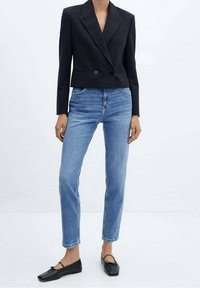 Blazer negro corto con solapas de pico, cierre de un solo botón, combinado con vaqueros azul claro de pierna recta y zapatos planos negros con detalle de lazo.