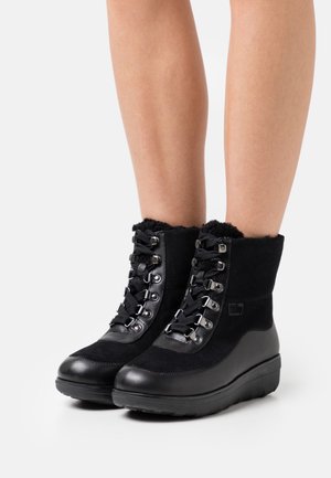 Bottines en cuir noir et daim avec lacets à l'avant et œillets métalliques, portées sur des jambes nues contre un fond blanc.