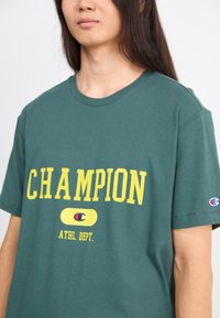 Zielona koszulka bawełniana z żółtym napisem "CHAMPION", okrągły dekolt, krótki rękaw i mały akcent z logo na rękawie.