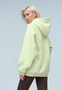 Haut à capuche oversize vert clair avec un cordon de serrage, des épaules tombantes et des poignets côtelés. Accompagné de leggings bruns côtelés.
