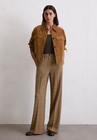 Braune Cord-Jacke mit zwei vorderen Taschen, kombiniert mit beigen Karo-Hosen mit hoher Taille. Das Modell steht mit den Händen in den Taschen und schaut nach vorne.