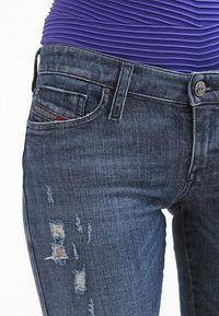 Jeans de mezclilla en azul oscuro con parches desgastados, un diseño ajustado, estilo de cinco bolsillos y un cierre de botón plateado en la cintura.