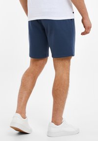 Shorts bleu marine avec une texture lisse et une taille élastique, présentant une coupe droite et des poches sur les côtés, associés à des baskets blanches.