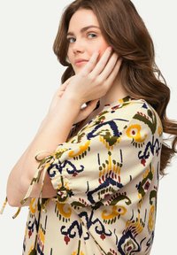Crème kleurige blouse met een levendig bloemen- en geometrisch patroon in marineblauw, geel, rood en groen, met gerimpelde mouwen en strikdetails.
