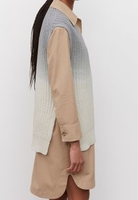 Veste en tricot dégradé du gris au crème, avec une partie supérieure texturée et une partie inférieure unie, portée sur une robe chemise beige à col avec des fentes latérales.