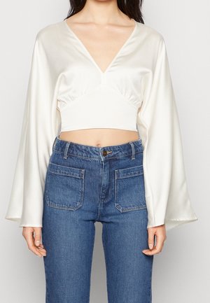 Femme portant un crop top en satin blanc à manches longues, col en V profond et manches larges, associé à un jean bleu taille haute avec des poches plaquées à l'avant.