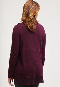 Langärmliger, tief burgunderfarbener Pullover aus weichem Material mit leicht asymmetrischem Saum und geschichtetem Rückendesign, ausgestattet mit einem runden Halsauschnitt.