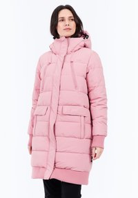 Mujer vestida con un abrigo de invierno acolchado y largo de color rosa, con capucha, bolsillos frontales y de pecho, de pie contra un fondo blanco.