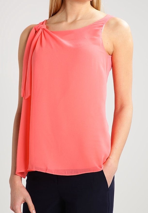 Blouse corail rose sans manches avec un détail de nœud sur une épaule, portée avec un pantalon sombre, modèle debout avec une main dans la poche.