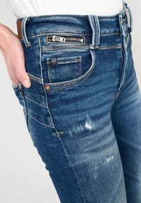 Slanke blauwe denim jeans met een verouderde afwerking, voorzien van een ritszak, messing accenten en zichtbare stikdetails.