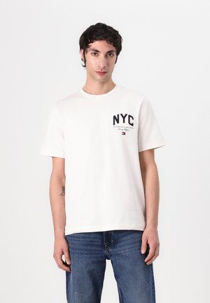Biela bavlnená tričko s námorníckym grafikom "NYC" na hrudi a tmavými akcentmi. Spárované s modrými džínsami. Krátke rukávy, okrúhly výstrih.