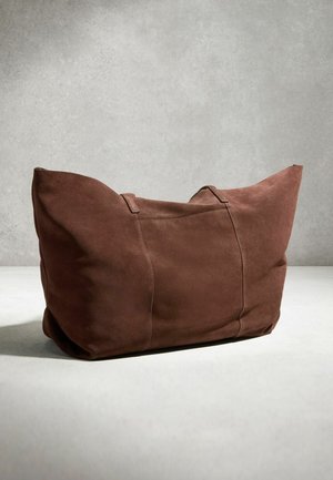 Große braune Wildledertasche mit zwei Griffen, die auf einer hellen Oberfläche vor einem schlichten grauen Hintergrund liegt.