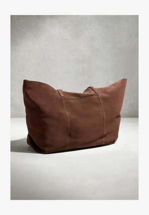 Große braune Wildledertasche mit zwei Griffen, die auf einer hellen Oberfläche vor einem schlichten grauen Hintergrund liegt.