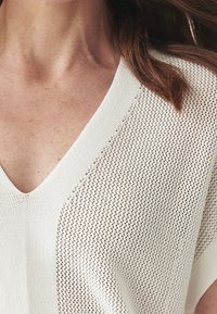 Detailní pohled na krk a rameno osoby v bílém texturovaném úpletu s výstřihem do V, s rozpuštěnými hnědými vlasy.