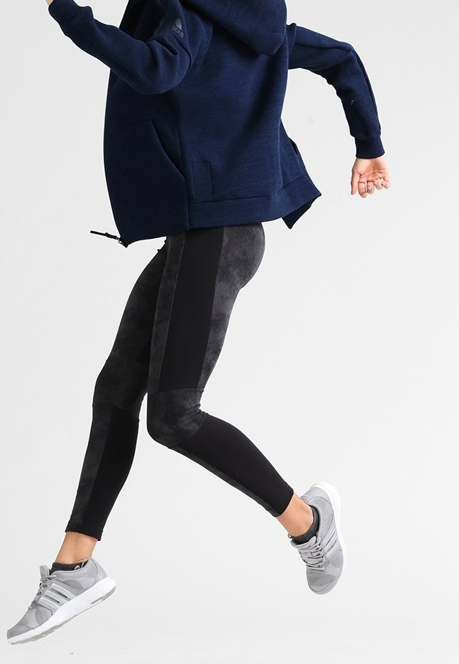 Personne en plein saut portant un sweat à capuche bleu marine, des leggings noirs à motifs avec des panneaux latéraux, et des baskets de sport gris clair sur un fond uni.