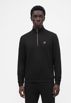 Junger Mann trägt einen schwarzen Langarmpullover mit Viertelreißverschluss und kleinem Logopatch auf der Brust vor neutralem Hintergrund.