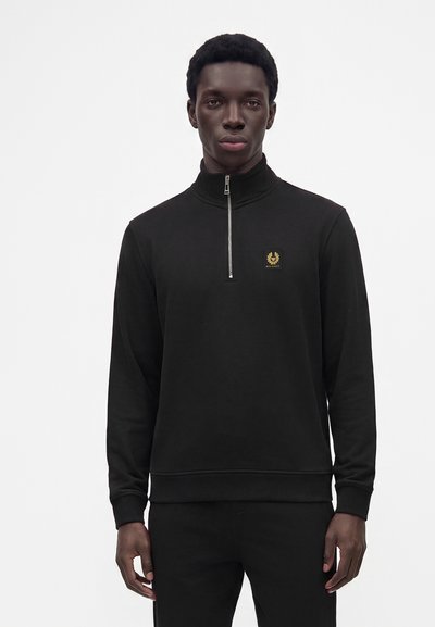Jeune homme portant un pull noir à manches longues avec une fermeture éclair en quart et un petit écusson logo sur la poitrine sur un fond uni.